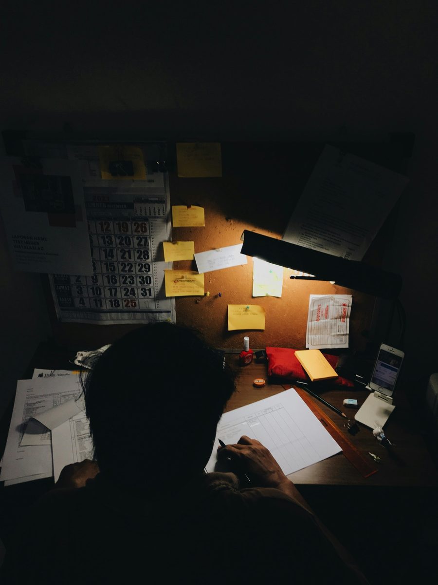 Una persona alla scrivania di notte, sotto la lampada, compila a mano un registro tra calendari e foglietti appesi